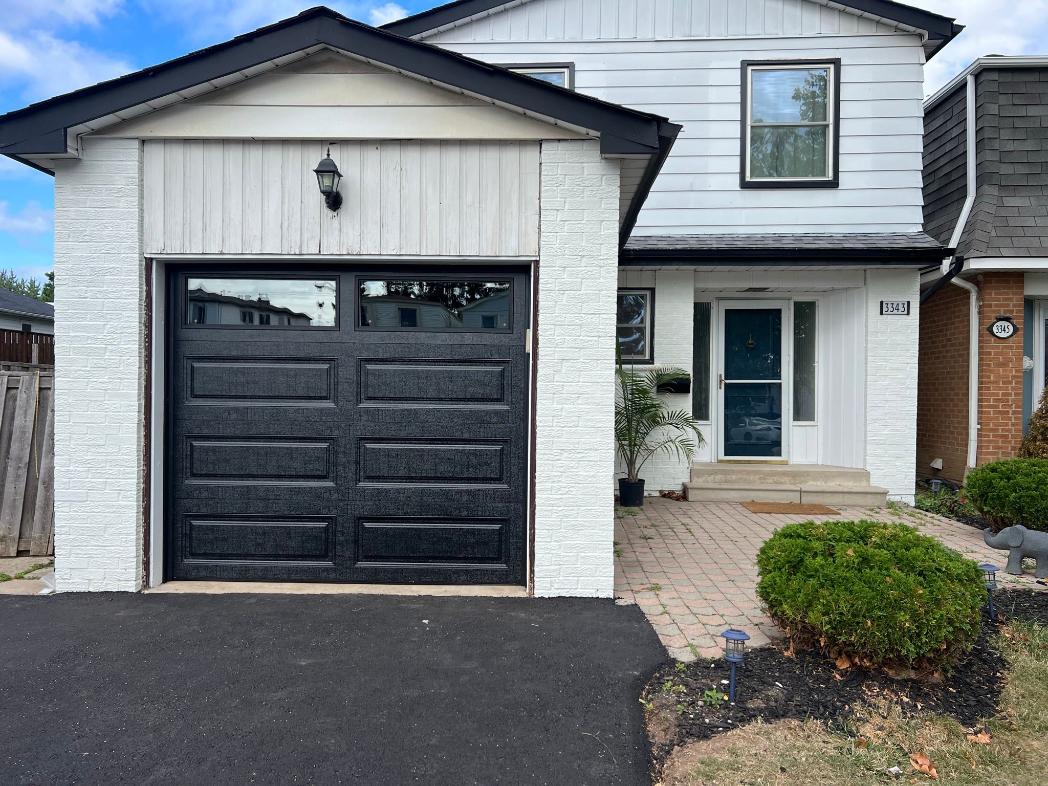 Black classic raised-panel garage door on a white and light-siding home with mature landscaping