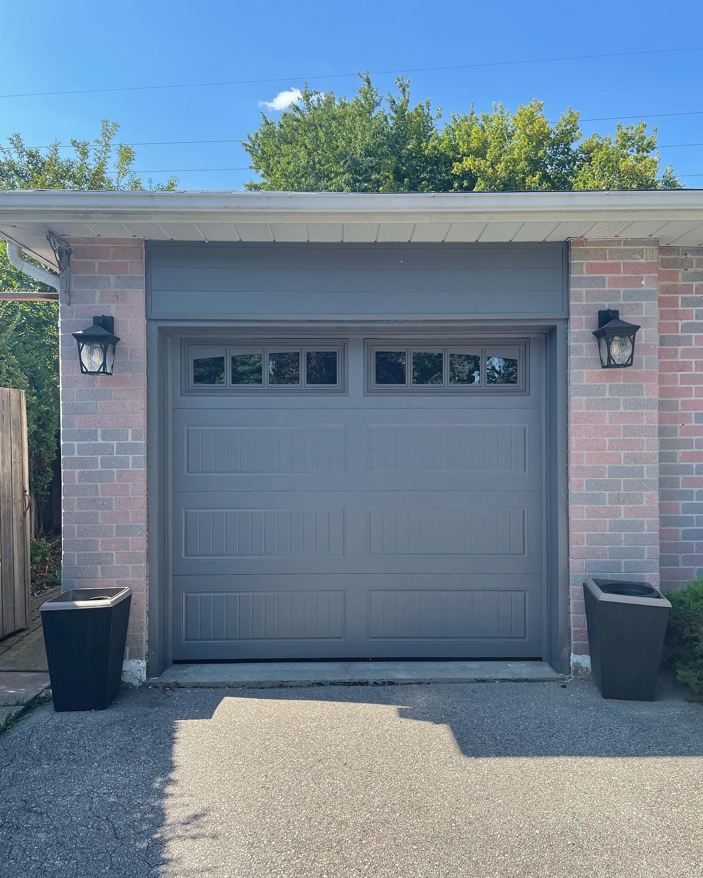 Grey traditional single-car garage door with top windows flanked by decorative planters on a sunny day