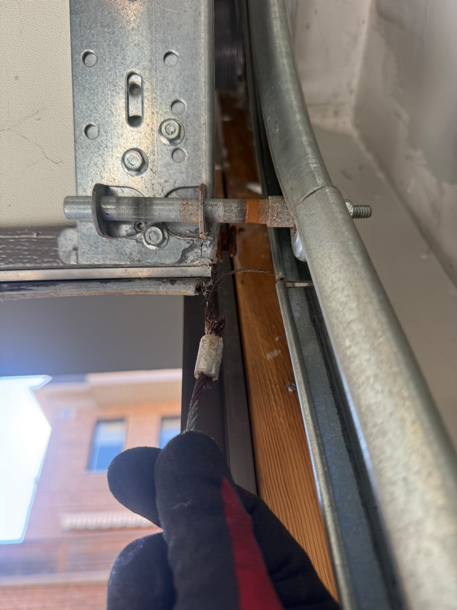 Gloved hand holding a snapped frayed garage door lift cable at a rusty bottom bracket roller - LTA field photo