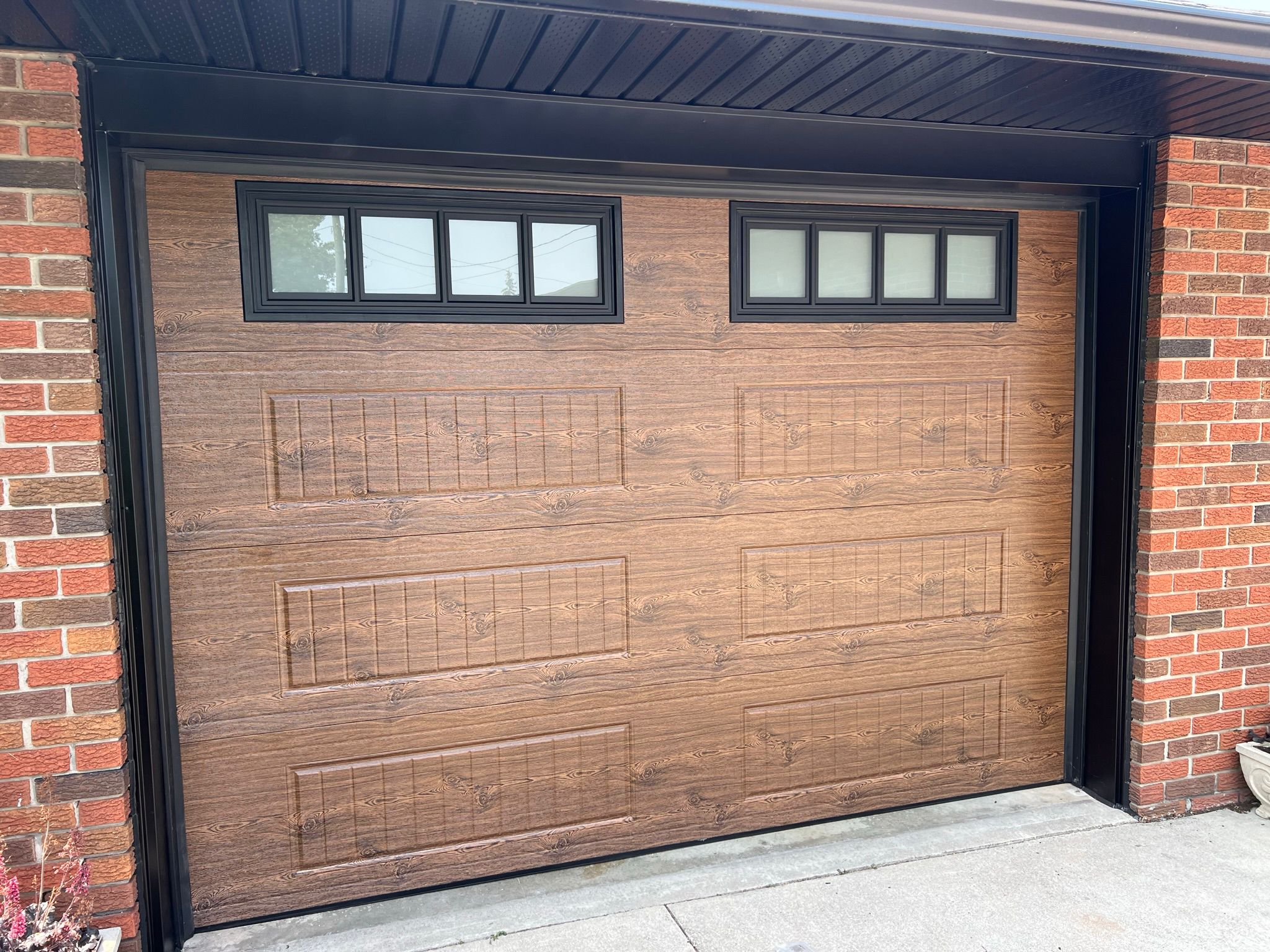 Medium walnut faux-wood single garage door with top windows on a red brick home surrounded by summer vegetation